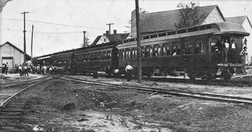 Historic black and white photo of a train station with a train and passengers gathered on the platform.