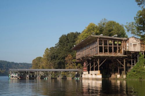 A rustic wooden structure on stilts by a calm lake, surrounded by trees and clear blue skies.