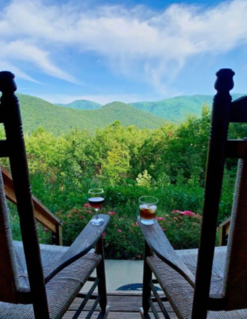 Two rocking chairs on a porch overlook a lush green landscape and distant mountains under a blue sky.