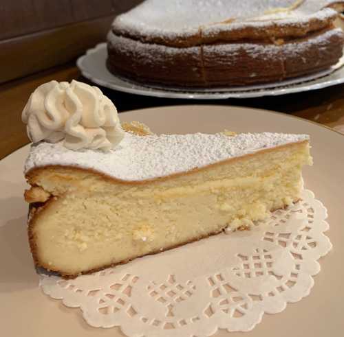 A slice of creamy cheesecake topped with whipped cream and dusted with powdered sugar, served on a lace doily.