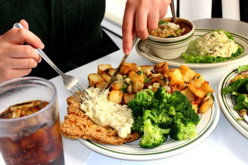 A person cuts into a plate of fried chicken topped with sauce, served with potatoes, broccoli, and a bowl of soup.