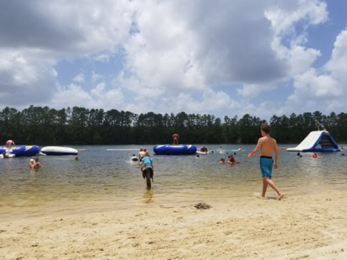 A sandy beach with people swimming and playing on inflatable structures in a lake under a partly cloudy sky.