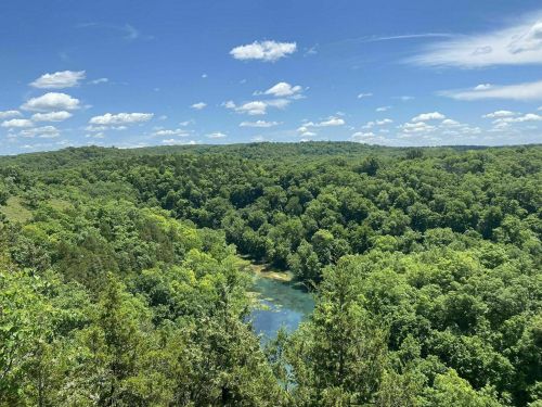 A lush green landscape with rolling hills, a clear blue sky, and a serene body of water surrounded by trees.