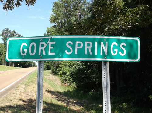 Green road sign reading "GORE SPRINGS" beside a rural road, surrounded by trees and clear blue sky.