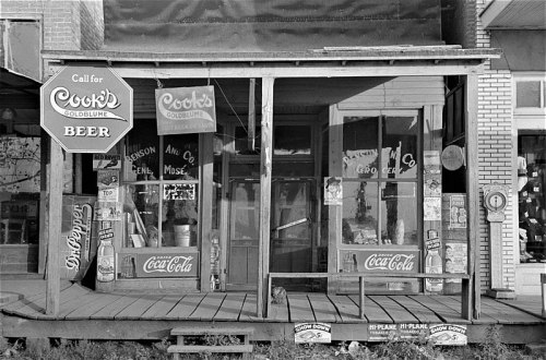 A vintage storefront with signs for Cook's Goldblume Beer and Coca-Cola, featuring a wooden porch and old advertisements.