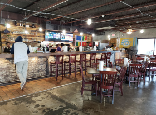 A casual restaurant interior with wooden tables, a bar area, and colorful flags hanging on the wall.