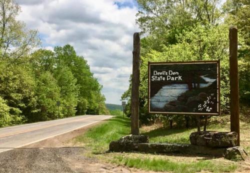 Sign for Devil's Den State Park beside a road, surrounded by lush green trees and a cloudy sky.