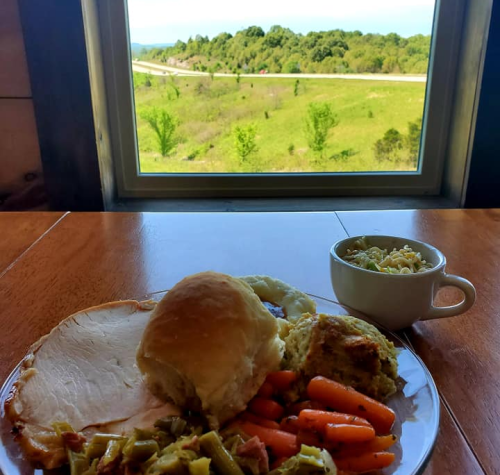 A plate of turkey, vegetables, and a roll sits on a table with a scenic view of green hills and a road outside.