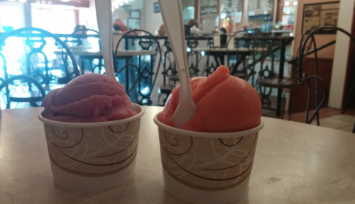 Two cups of colorful sorbet, one pink and one orange, with white spoons, on a table in a café.