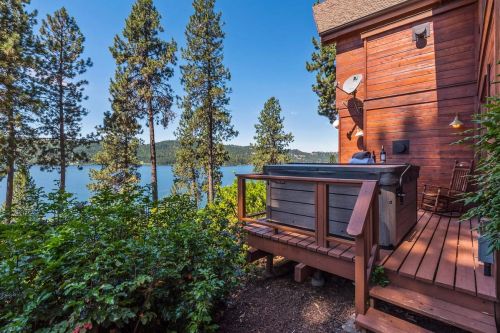 A wooden deck with a hot tub overlooks a serene lake surrounded by tall pine trees and mountains in the background.