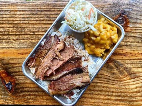 A metal tray with sliced brisket, rice, macaroni and cheese, and a side of coleslaw on a wooden table.