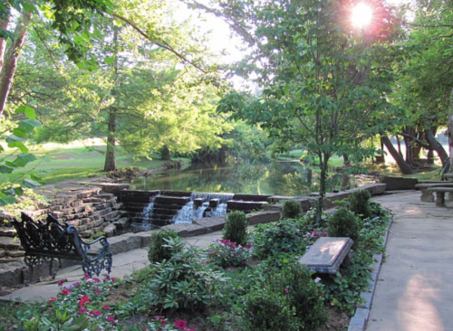A serene park scene featuring a small waterfall, lush greenery, and a bench beside a tranquil pond.