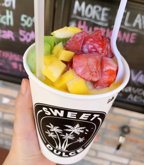 A hand holding a cup of fruit topped with mango, strawberries, and green fruit, with a "Sweet Dulce" logo.