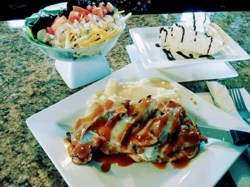 A plate with grilled chicken topped with sauce, mashed potatoes, a side salad, and a slice of dessert on a table.