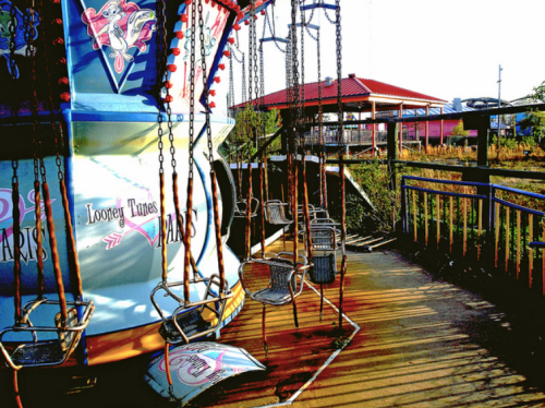 Colorful amusement park ride with empty swings, surrounded by vibrant scenery and a red-roofed building in the background.