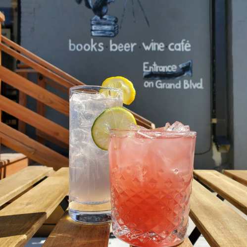 Two cocktails on a wooden table, with a wall in the background featuring a sign for a books, beer, and wine café.