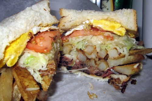 A close-up of a hearty sandwich filled with layers of meat, lettuce, tomato, fried egg, and crispy fries between two slices of bread.