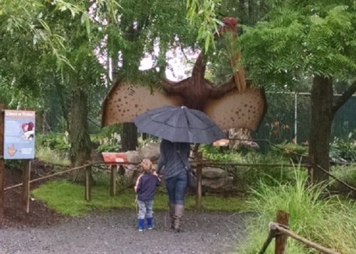 A person and a child stand under an umbrella, facing a large dinosaur sculpture in a park.