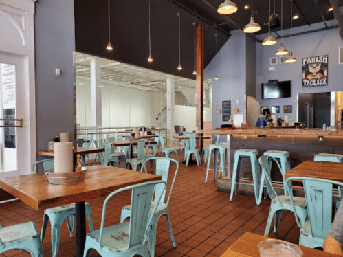 A modern restaurant interior with wooden tables, turquoise metal chairs, and a bar area in the background.