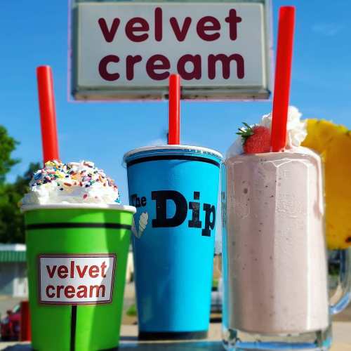 Three colorful drinks with whipped cream and toppings, featuring a green cup, a blue cup, and a tall glass, against a sign.