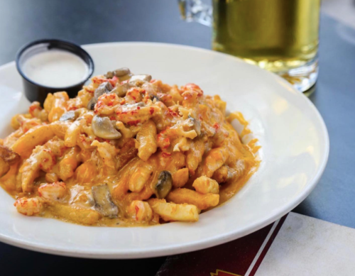 A plate of creamy pasta with seafood, topped with mushrooms, served with a side of dipping sauce and a glass of beer.