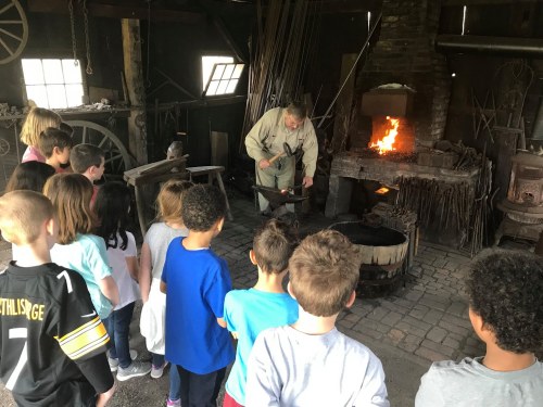 A group of children watches a blacksmith demonstrate his craft in a historic workshop with a fire in the forge.