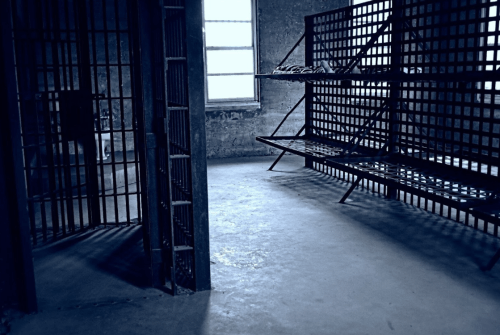 A dimly lit prison cell with metal bars and empty bunk beds against a concrete wall.