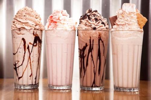 Four tall glasses of milkshakes topped with whipped cream, chocolate syrup, and various toppings, arranged on a wooden table.