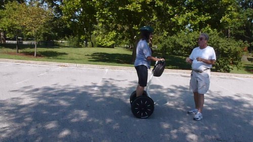 A person on a Segway talks to another person in a park with trees in the background.