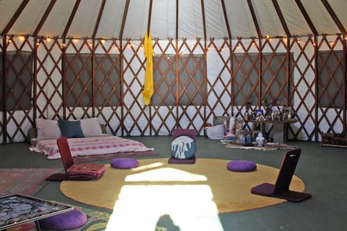 Interior of a yurt with a circular rug, seating cushions, and decorative items on shelves. Natural light filters in.