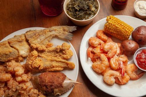 A plate of fried fish, shrimp, corn, potatoes, and greens, served with dipping sauce on a wooden table.