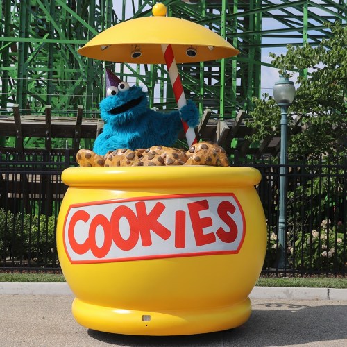 A blue, furry character with a cookie jar hat holds a straw, surrounded by cookies in a large yellow jar labeled "COOKIES."