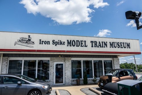 Exterior of the Iron Spike Model Train Museum, featuring a sign and parked cars under a blue sky with clouds.