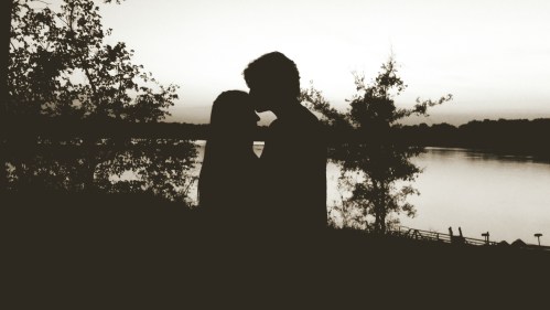 Silhouette of a couple embracing by a lake at sunset, surrounded by trees.