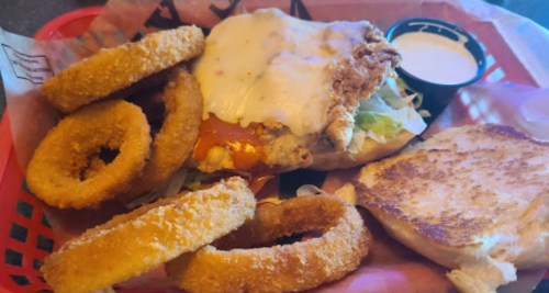 A basket with a fried chicken sandwich topped with cheese, lettuce, and tomato, accompanied by onion rings and a dipping sauce.