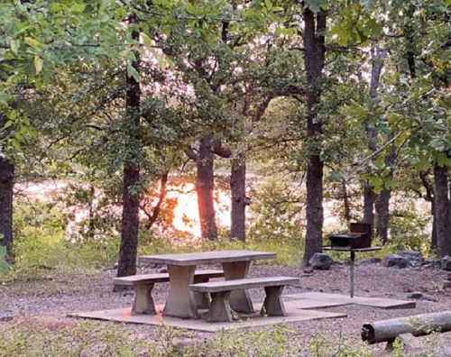 A picnic table and grill surrounded by trees, with a sunset reflecting on a nearby body of water.