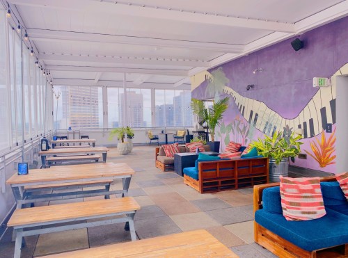 A bright rooftop lounge with wooden tables, cozy seating, and a colorful mural of piano keys on the wall.
