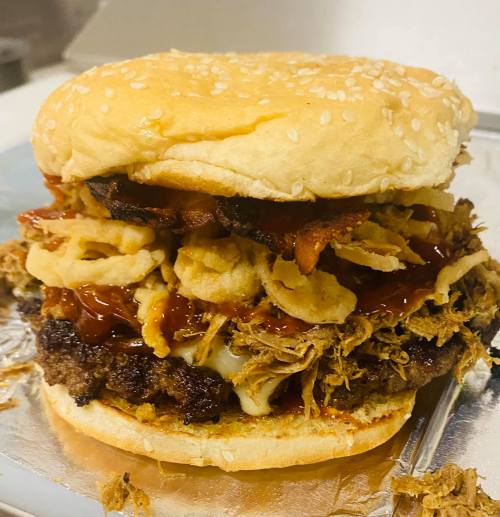 A towering burger with a sesame seed bun, beef patty, pulled pork, crispy onions, and barbecue sauce.