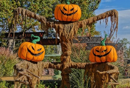 Three carved pumpkins with spooky faces are stacked on a scarecrow-like structure, surrounded by autumn foliage.