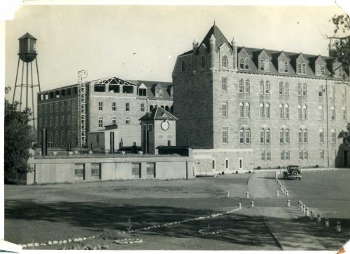 Historic building with stone architecture, a water tower, and a vintage car parked nearby. Black and white photo.