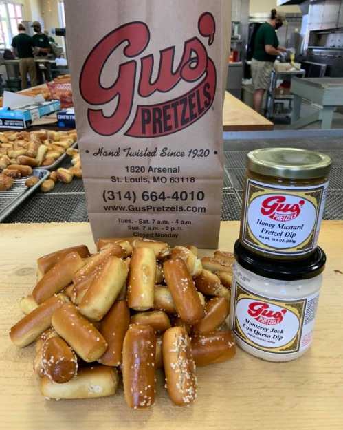 A pile of pretzel bites in front of a bag of Gus' Pretzels and jars of honey mustard and cheese dip.