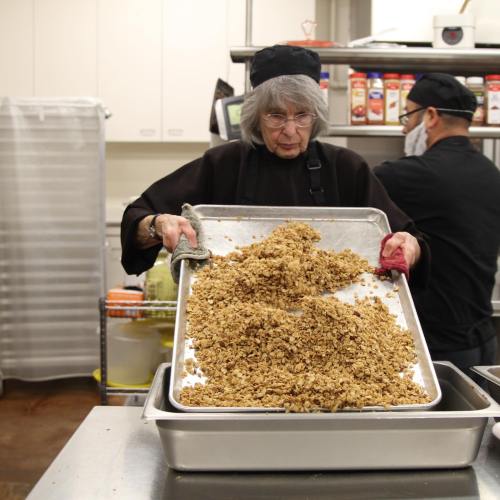 A person in a kitchen wearing a black hat and apron pours granola from a tray into a container.