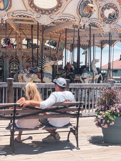 A man and a girl sit on a bench, watching a carousel with colorful horses in a sunny outdoor setting.