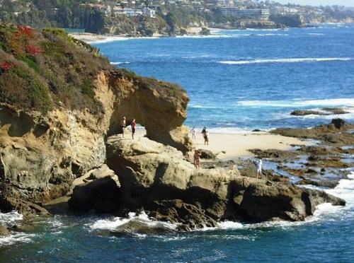 People exploring rocky cliffs by the ocean, with a sandy beach and coastal homes in the background.