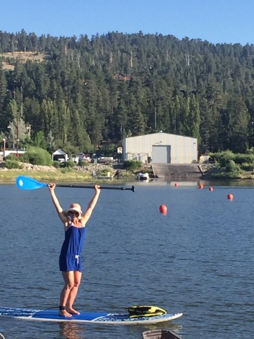 A person in a blue outfit stands on a paddleboard, raising their arms in celebration on a calm lake surrounded by trees.