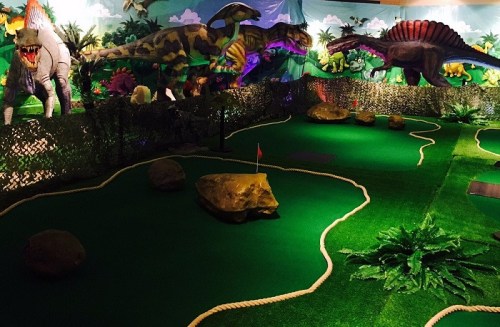 A mini-golf course featuring dinosaur decorations, green turf, rocks, and vibrant jungle-themed backgrounds.