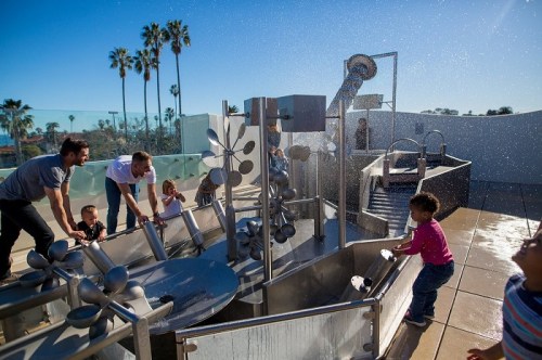 Children and adults play with water features at a sunny outdoor park, surrounded by palm trees.
