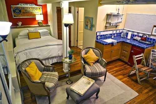 Cozy studio apartment with a bed, kitchenette, and seating area featuring yellow accents and wooden floors.