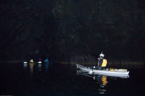 A kayaker in a dark cave, with two other kayakers visible in the background, reflecting on the calm water.