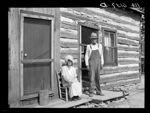 A man in overalls stands beside a woman in a dress and bonnet, sitting on a porch of a log cabin.